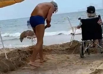 Joven remueve la arena de la playa para que su mamá en silla de ruedas sienta el mar en los pies