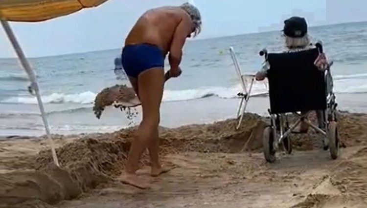 Joven remueve la arena de la playa para que su mamá en silla de ruedas sienta el mar en los pies